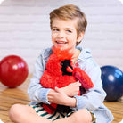 A young child with a wide, happy smile holding the Red Cardinal by Warmies plush toy while seated against a neutral background with colorful balloons nearby.