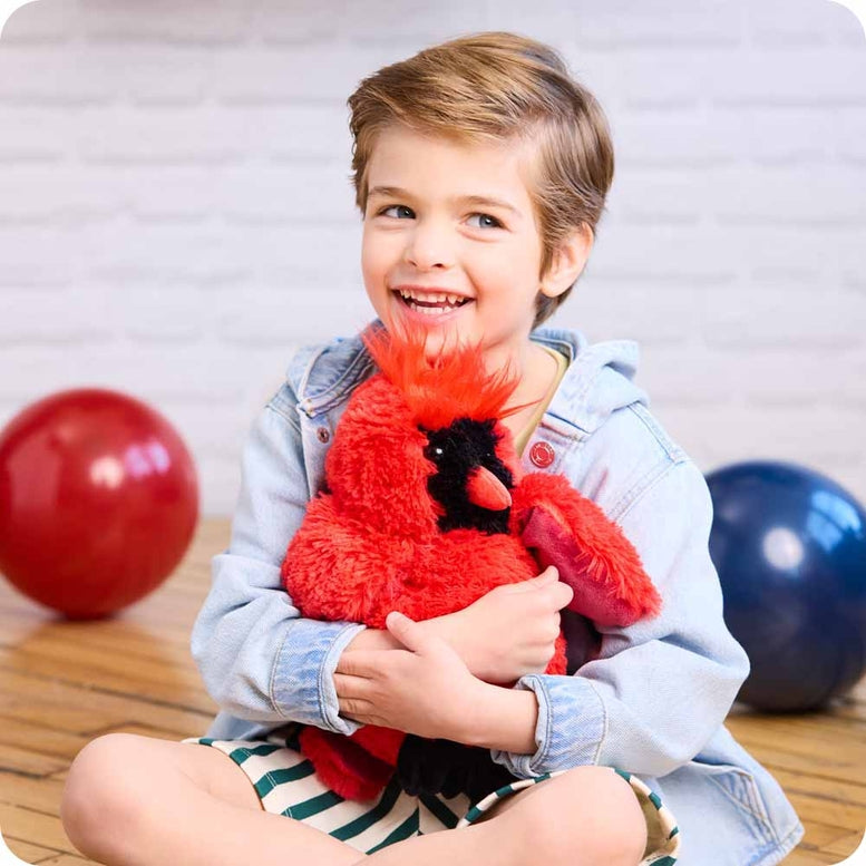 A young child with a wide, happy smile holding the Red Cardinal by Warmies plush toy while seated against a neutral background with colorful balloons nearby.