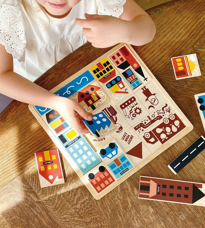 A young girl with blonde hair tied back sits at a wooden table, smiling as she plays with the Mentari Wooden Town Puzzle, holding a puzzle piece in her hand while assembling the colorful town scene.