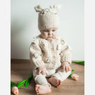 Baby wearing a cream knitted outfit with a matching hat on a wooden floor.