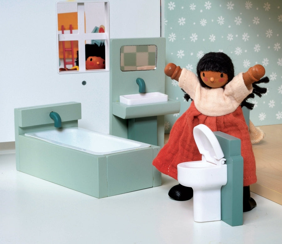 A lifestyle product photo of the Wooden Bathroom Furniture set by Mentari set up in a miniature dollhouse bathroom, featuring a doll with dark hair and a red dress standing near the toilet and sink.