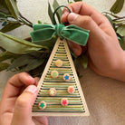 Handmade wooden Christmas tree ornament with colorful buttons and a green ribbon, held by hands against a natural background.