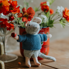 Knitted mouse toy in a blue sweater holding flowers on a wooden surface with floral background