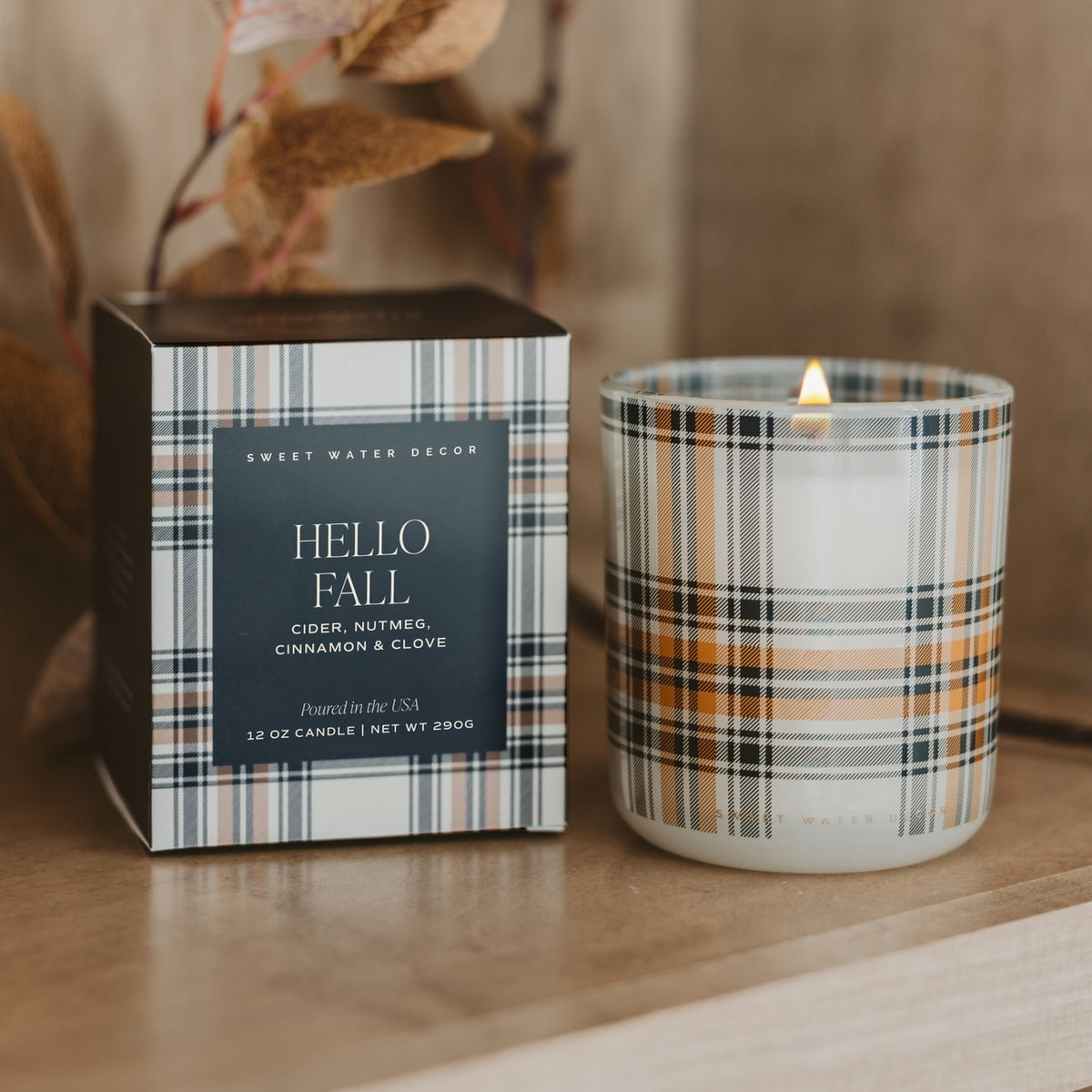A lit candle in a clear glass jar with a plaid pattern sits beside its matching box on a wooden surface. The packaging and glass feature a plaid design in navy, beige, white, and orange-brown tones, with a dark navy center label on the box reading 'HELLO FALL' and 'SWEET WATER DECOR'.