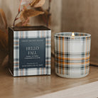 A lit candle in a clear glass jar with a plaid pattern sits beside its matching box on a wooden surface. The packaging and glass feature a plaid design in navy, beige, white, and orange-brown tones, with a dark navy center label on the box reading 'HELLO FALL' and 'SWEET WATER DECOR'.