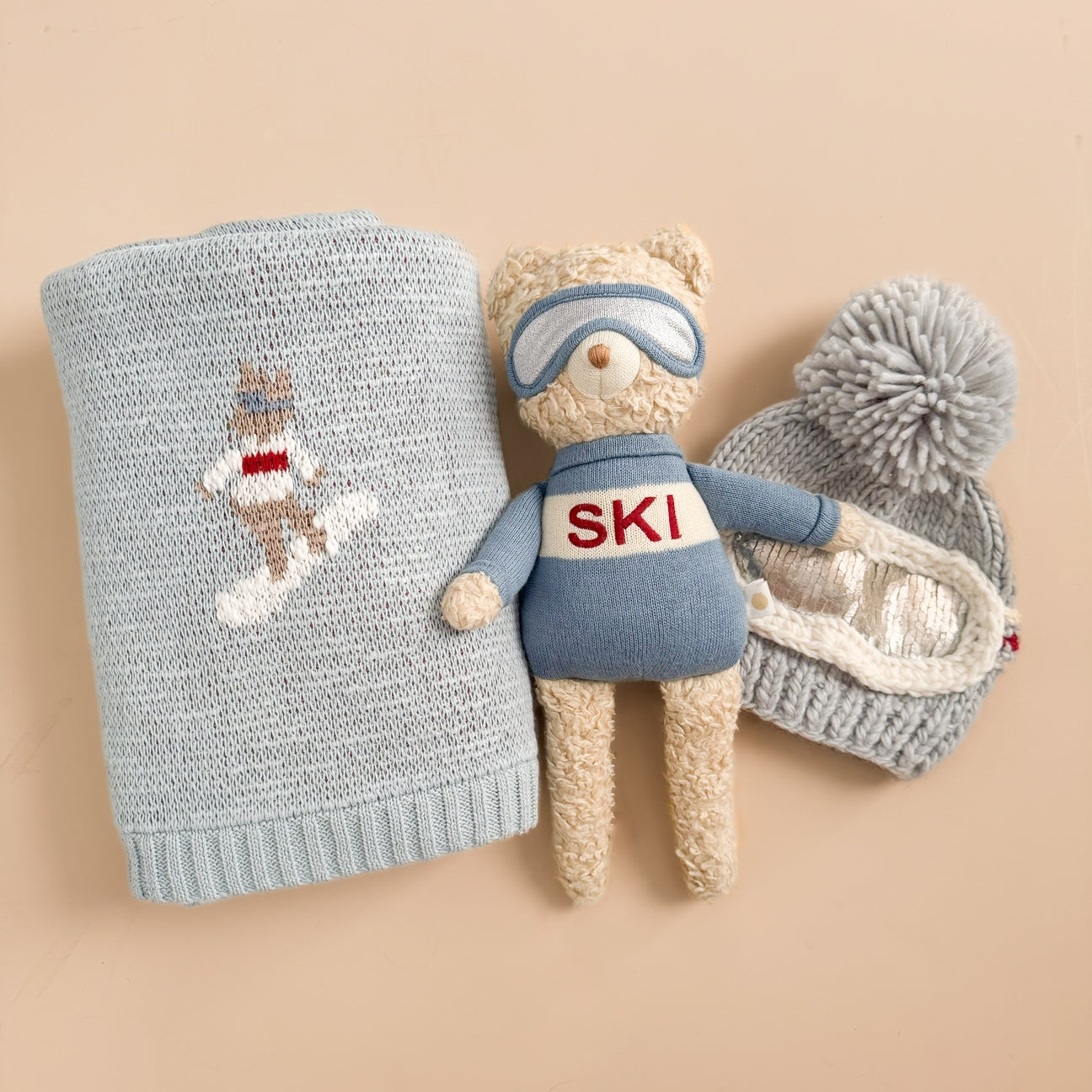 Knitted blanket with snowman design, teddy bear toy with 'SKI' sweater, and gray knit hat on beige background