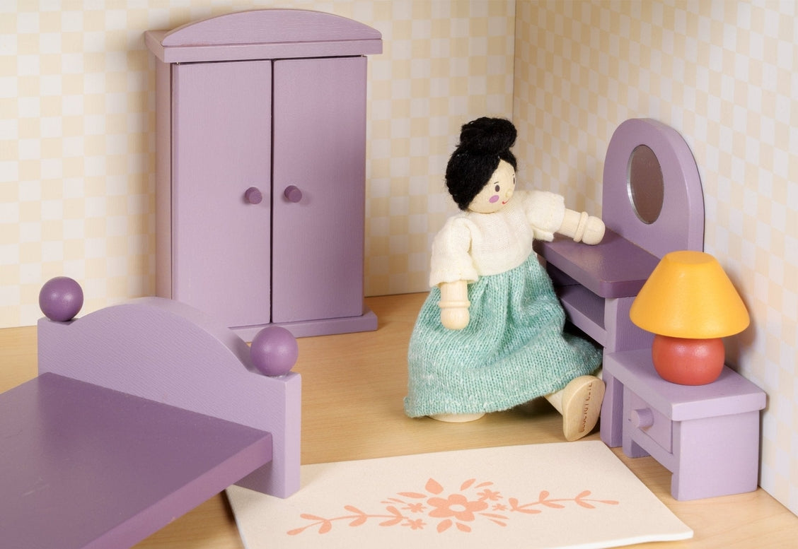 A dollhouse bedroom scene featuring a wooden doll in a green dress sitting on the edge of a double bed with a purple frame and bedding. Against the back wall stands a purple wooden wardrobe with closed doors, flanked by a dressing table with an oval mirror and a matching stool. Next to the bed is a bedside table topped with a lamp featuring an orange base and a yellow shade, and a small floral-patterned rug rests on the floor in the foreground.
