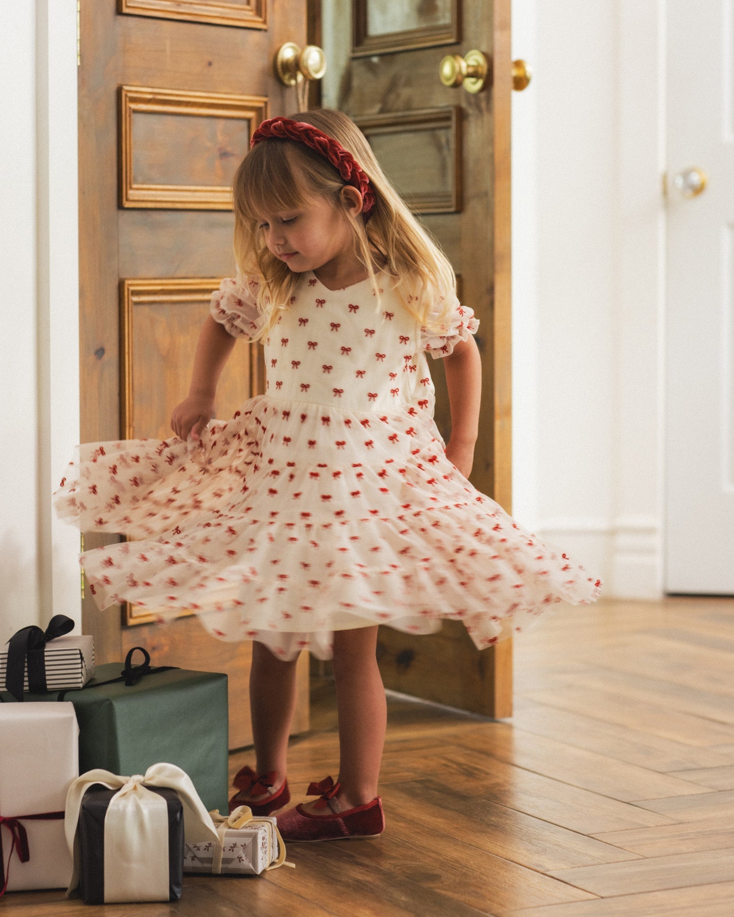 A young girl with blonde hair and a red headband twirls in a white tulle dress with red embroidered stars, standing in a doorway with wooden floors and gift boxes at her side.