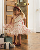 A young girl with blonde hair and a red headband twirls in a white tulle dress with red embroidered stars, standing in a doorway with wooden floors and gift boxes at her side.