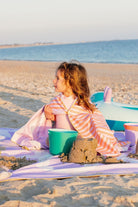 Child sitting on a beach with sandcastle and buckets, wrapped in a colorful towel.
