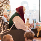 Person in a red and green outfit reading to children in a cozy room with Christmas decorations.