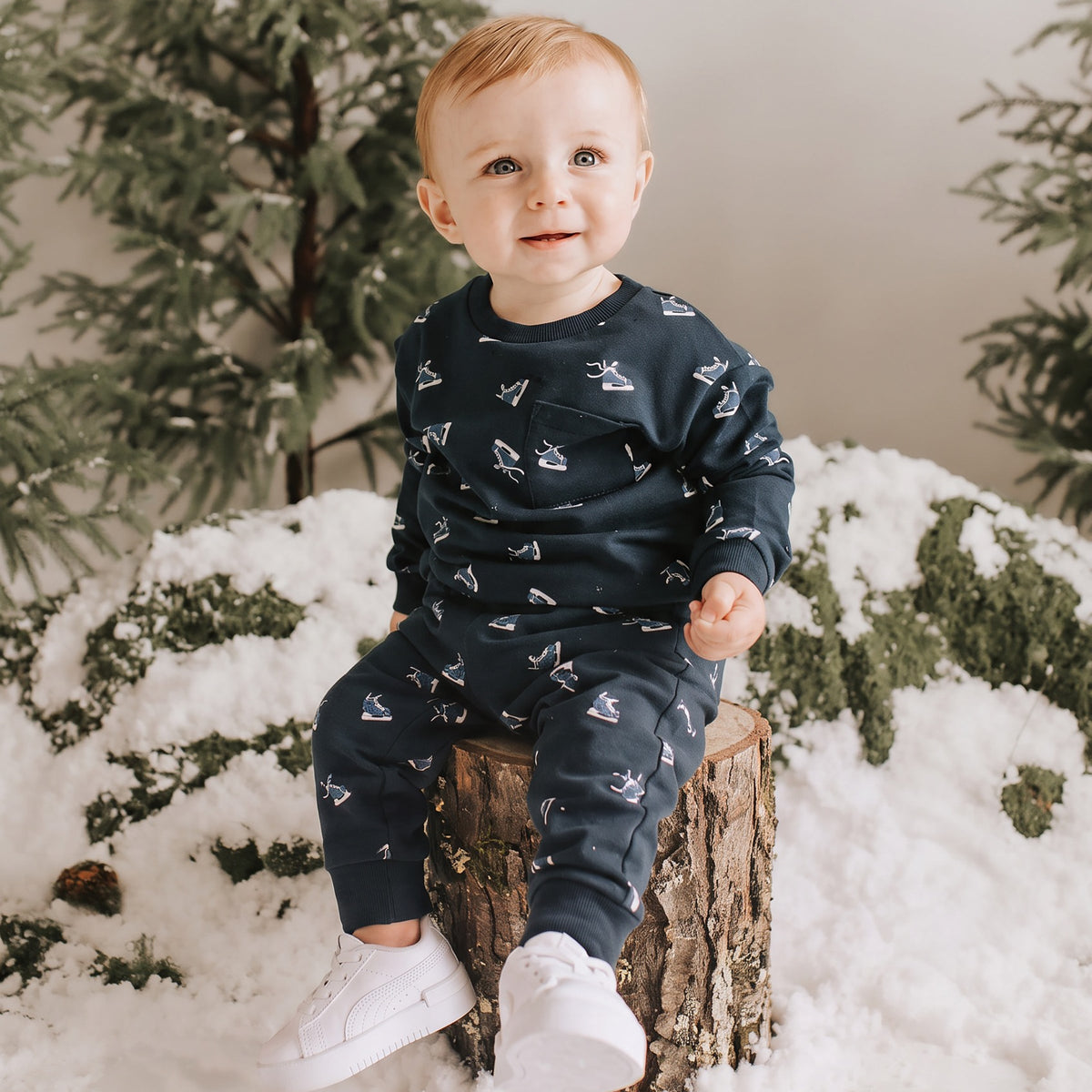 A child wearing two-piece kids' clothing set featuring a navy blue sweatshirt and matching jogger pants, both decorated with a repeating pattern of small white and light blue ice skaters, on a tree and snow white background.