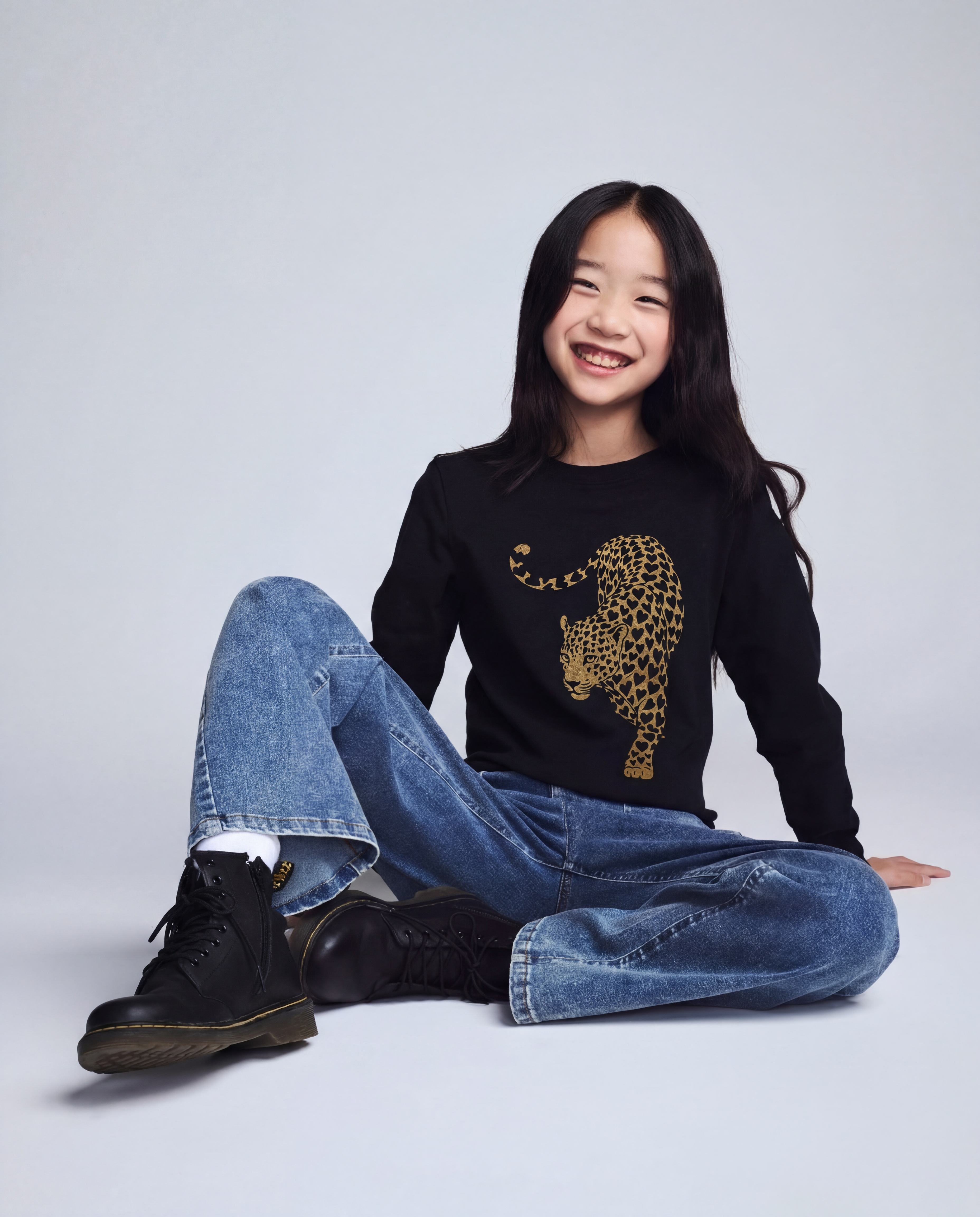 Person wearing a black sweater with a leopard print design, blue jeans, and black boots on a light gray background