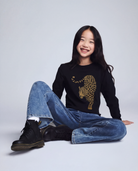 Person wearing a black sweater with a leopard print design, blue jeans, and black boots on a light gray background
