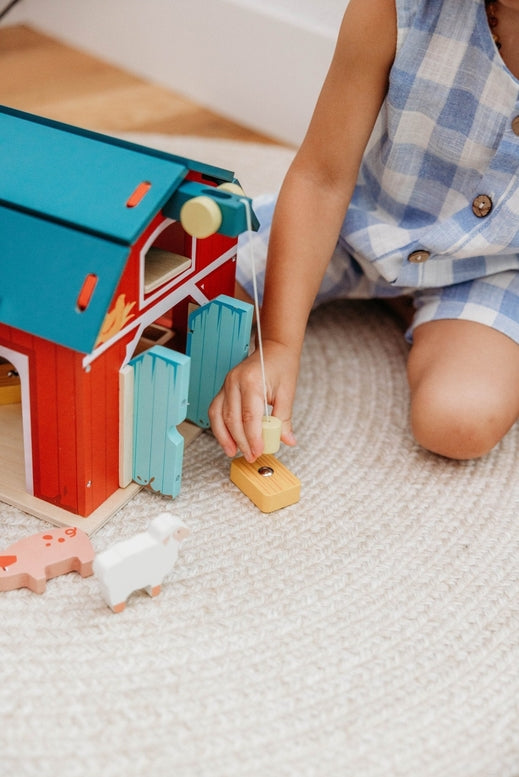 A child’s hands operating the working pulley system on the Wooden Big Red Barn Playset by Mentari, lifting a yellow magnetic hay bale toward the loft while open double doors reveal a glimpse inside the red barn.