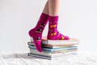 A person's legs wearing bright pink crew socks with a playful cat and dog pattern, standing on a stack of books on a white textured blanket against a white background.