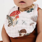 A close-up of a baby wearing a white muslin bib with a colorful printed design of red and brown mushrooms, green leaves, and a dragonfly, paired with a brown ribbed knit top.