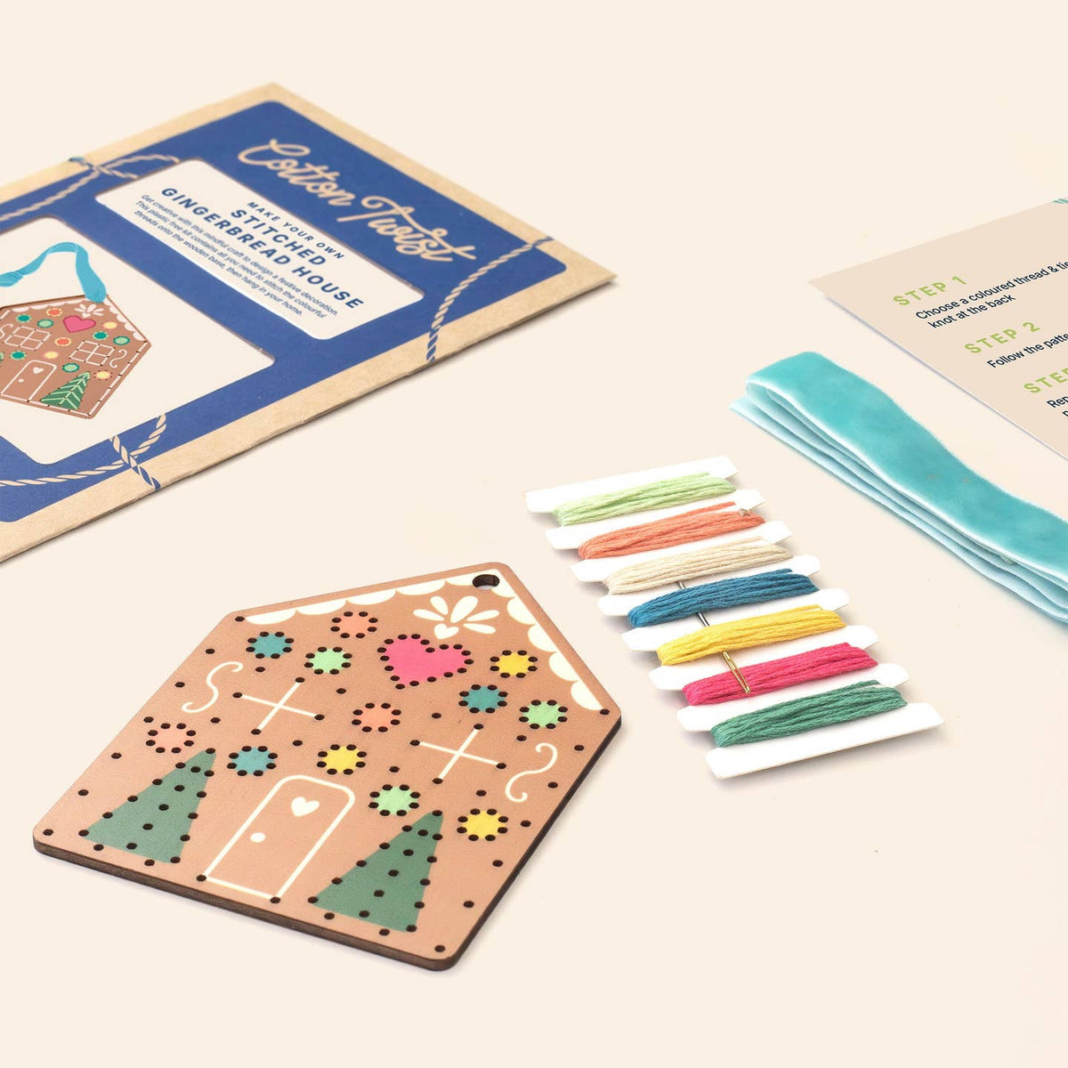 A gingerbread house embroidery kit laid out on a light beige background, including a wooden house-shaped embroidery board, a blue instruction booklet, a card with step-by-step instructions, colorful thread spools, and teal ribbon.