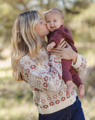 A woman in a cream knit sweater with a rust-brown floral pattern holds and kisses a baby wearing a matching dark burgundy outfit and a small bow headband, set against a softly blurred outdoor background.