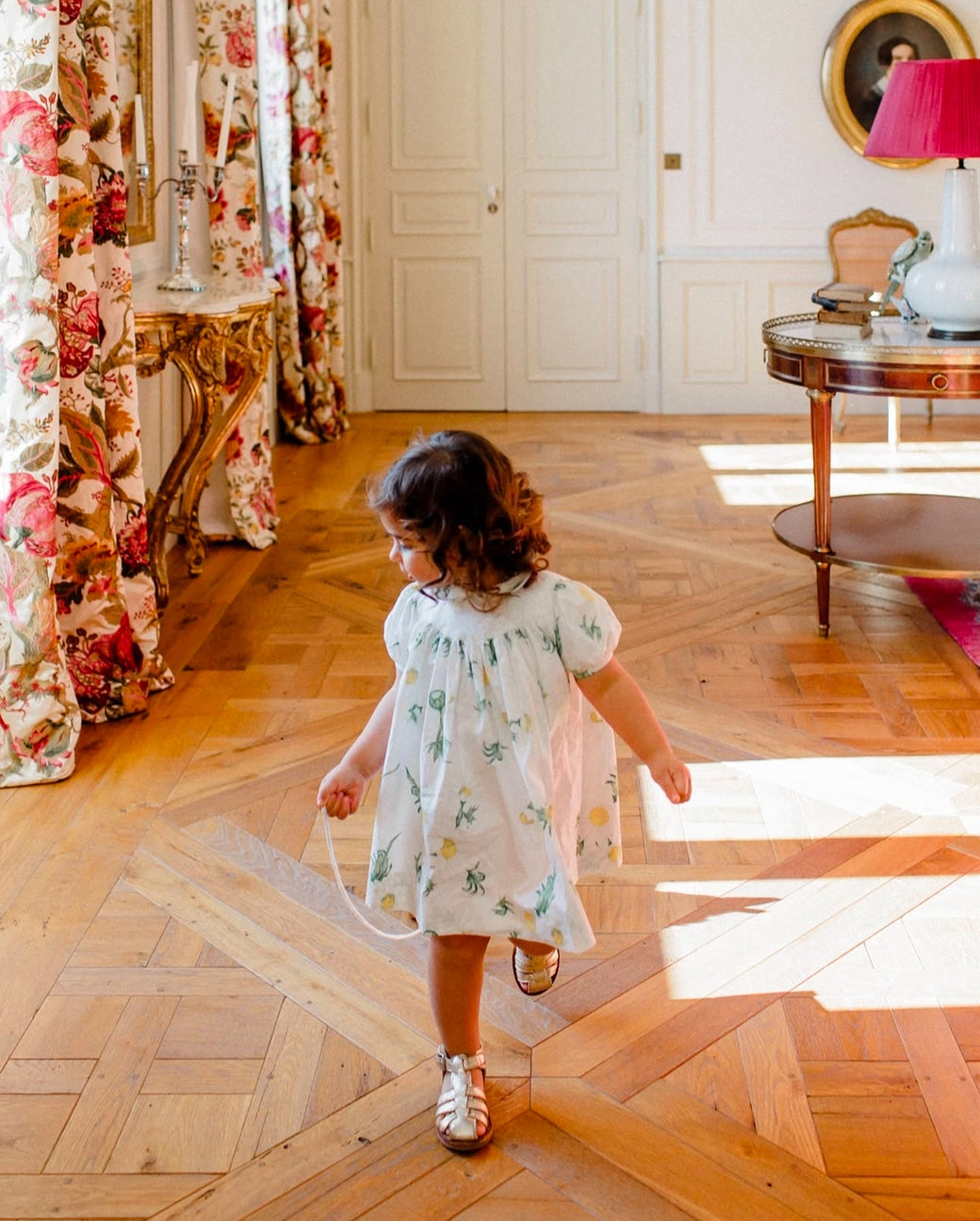 Young girl in a floral dress walking on a wooden floor in an elegant room with floral curtains and a lamp.