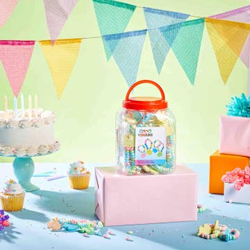 A clear plastic jar filled with colorful lollipops and candy sits on a pink gift box in a decorated party setting with pastel bunting, cupcakes, a cake, and wrapped presents.