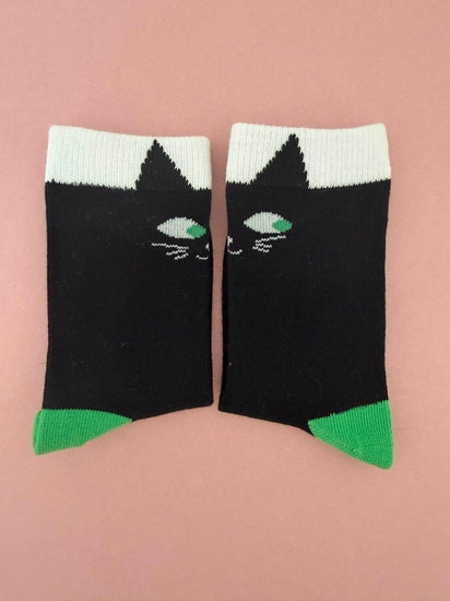 A pair of black crew socks with a white cuff, featuring a stylized cat face design with green eyes and a green toe cap, displayed on a solid pink background.