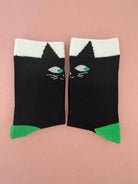 A pair of black crew socks with a white cuff, featuring a stylized cat face design with green eyes and a green toe cap, displayed on a solid pink background.