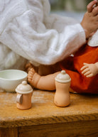 Two small wooden toy bottles with beige nipples and lids, displayed on a rustic wooden table next to a white bowl, with a person in a white sweater holding a doll in the background.