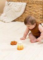 A baby in a brown sleeveless outfit crawls on a cream textured rug, playing with wooden stacking toys. A woven basket, a white knit blanket, and a soft beige background complete the cozy nursery setting.
