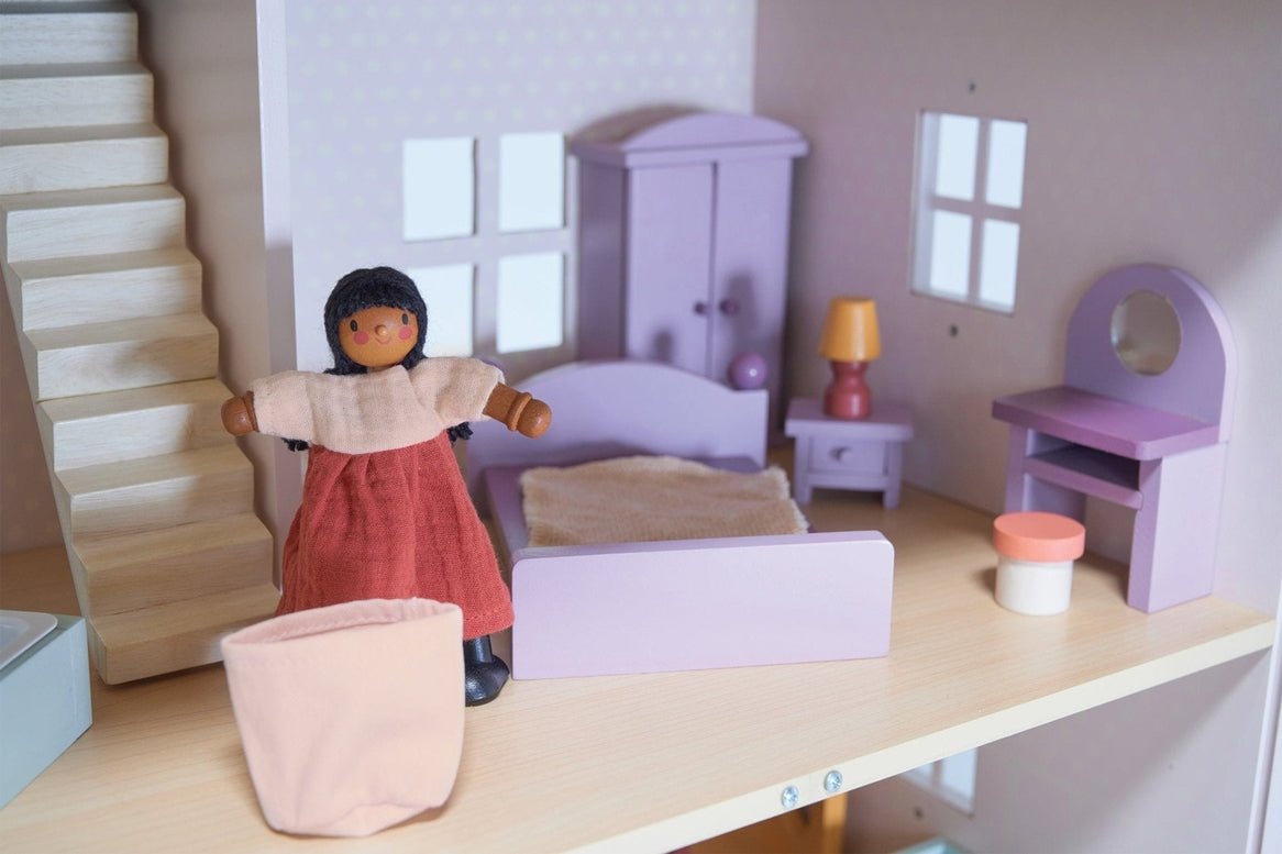 A dollhouse bedroom scene featuring a wooden doll in a reddish-brown dress standing near a white toy laundry basket in the foreground. In the background, a double bed with a purple frame and bedding sits against the wall, alongside a matching purple wardrobe, a bedside table with a yellow-shaded lamp, and a dressing table with a stool near a window.