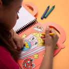 Child coloring a cartoon character in a spiral-bound coloring book with crayons on an orange background