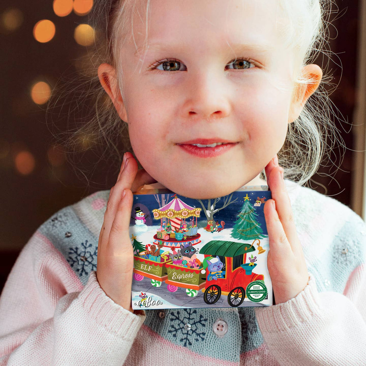 A young child with light hair holds a colorful illustrated Advent calendar box in front of their face. The box features a winter scene with a red train, Christmas tree, carousel, and gift boxes, with the text 'Advent Calendar' and 'Kreola' visible.