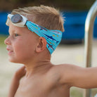 Young boy wearing a blue swim cap with shark pattern and goggles on his head, standing by a pool.