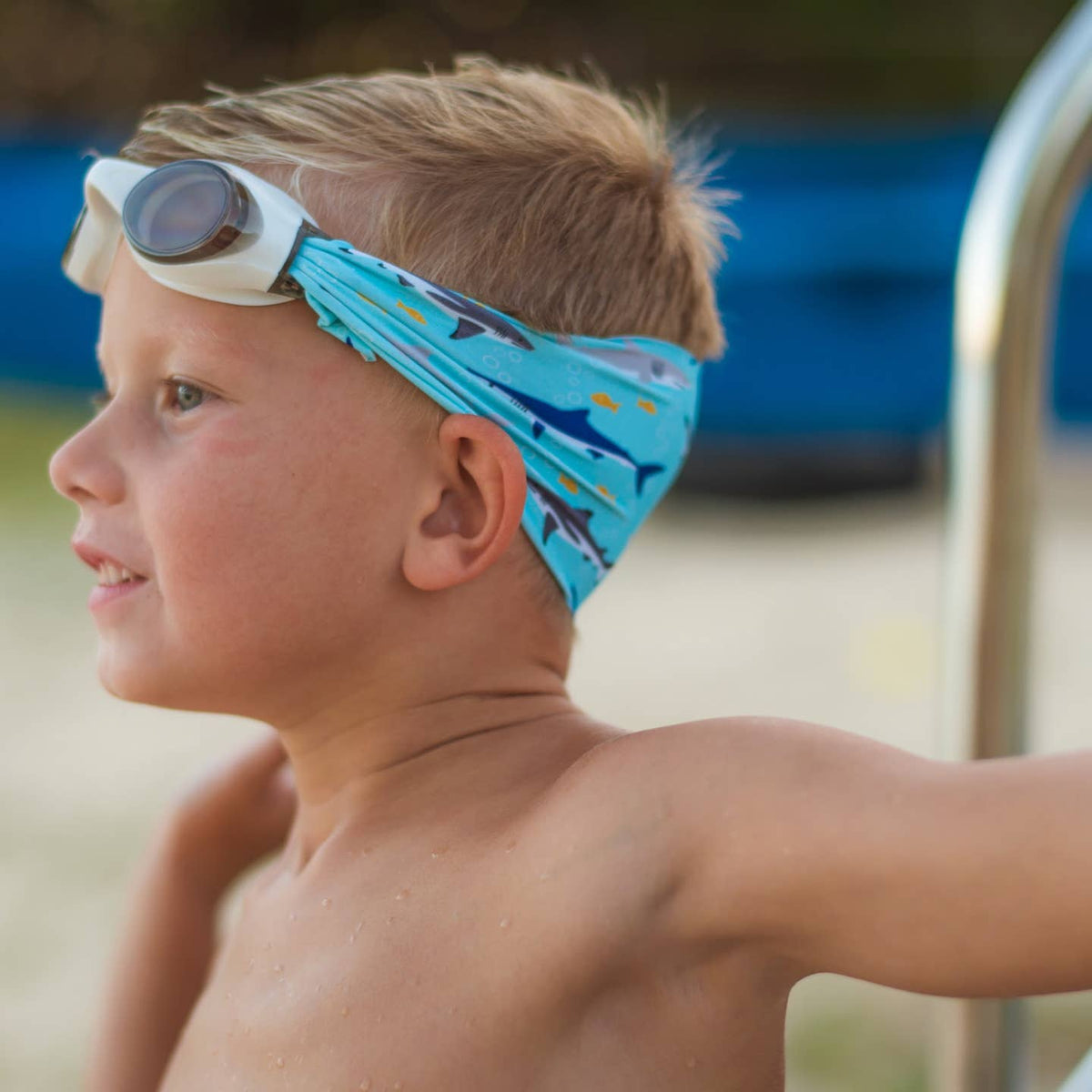 Young boy wearing a blue swim cap with shark pattern and goggles on his head, standing by a pool.