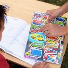 Child coloring with car-themed stickers on a book at a wooden table.