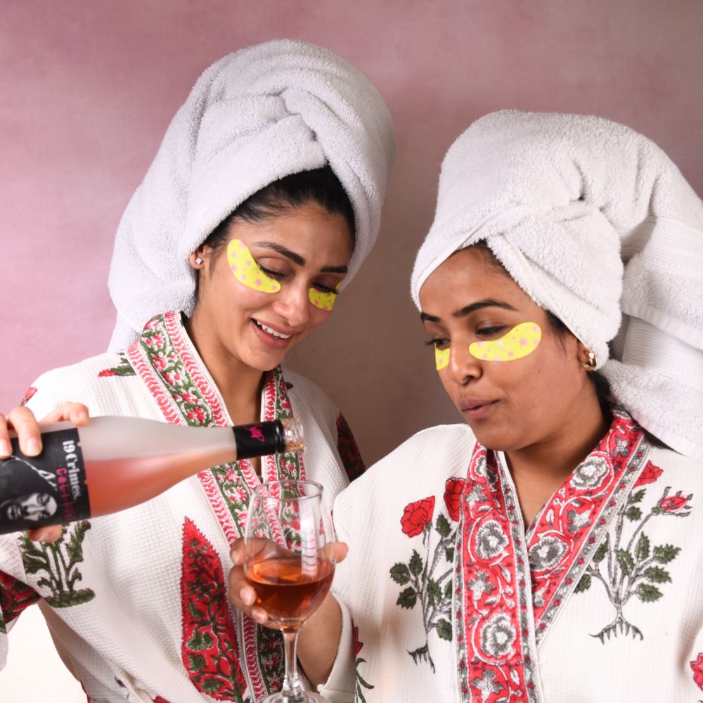 Two women in bathrobes with towels on heads, one pouring a drink and the other holding a glass, against a pink background.