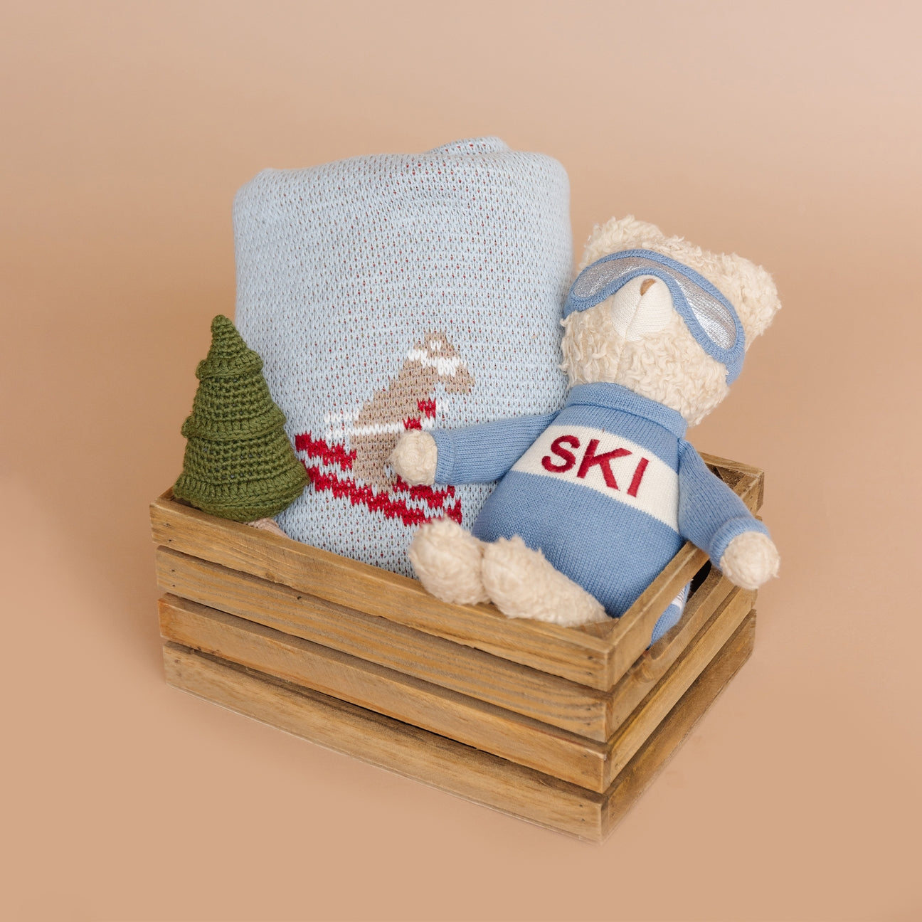 Teddy bear in a 'SKI' outfit with a knitted blanket and tree on a wooden crate against a beige background