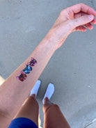 A temporary tattoo of three stacked cat heads with colorful glitter-style eyes and pink ears, applied on a person's forearm. The photo is taken outdoors on a gray concrete surface, with the person's legs and white socks visible below.