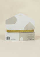 A house-shaped white display card holding a single strip of glittery gold ribbon, photographed against a plain warm beige background.
