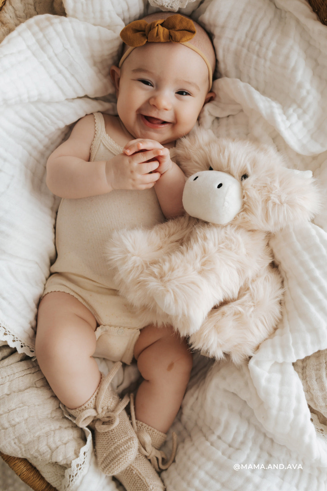 Baby lying on a soft surface with a fluffy white teddy bear, wearing a beige romper and headband.