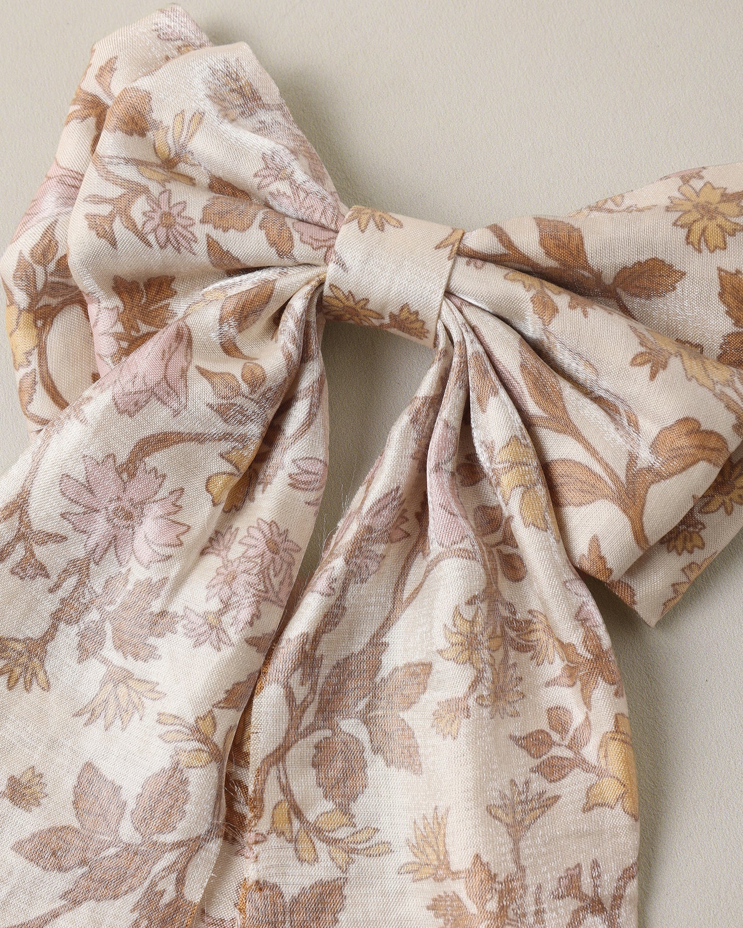 A large fabric bow featuring a delicate floral and leaf pattern in muted brown and dusty pink on a warm cream background, photographed against a neutral beige wall.
