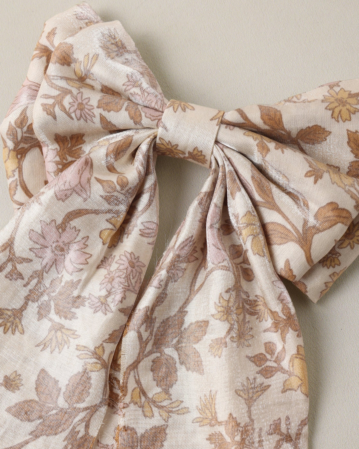 A large fabric bow featuring a delicate floral and leaf pattern in muted brown and dusty pink on a warm cream background, photographed against a neutral beige wall.