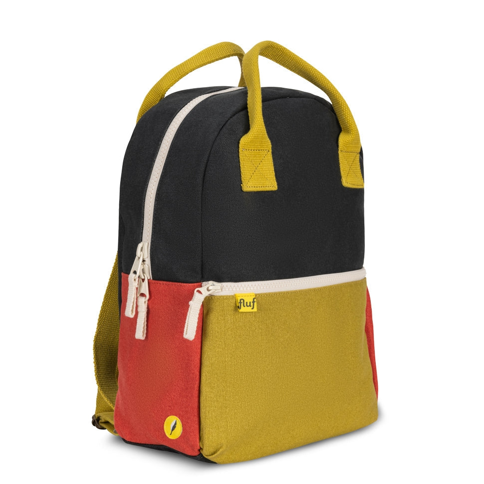 A color-blocked backpack by Fluf, featuring a black top half, a red front pocket, and a mustard yellow lower half, with mustard yellow handles and white zipper pulls, shown on a plain white background.