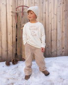 A young child wearing a cream knit beanie, a cream sweatshirt with 'HOT COCOCA CLUB' text, and beige-and-white vertically striped pants, standing outdoors in snow in front of a wooden fence.