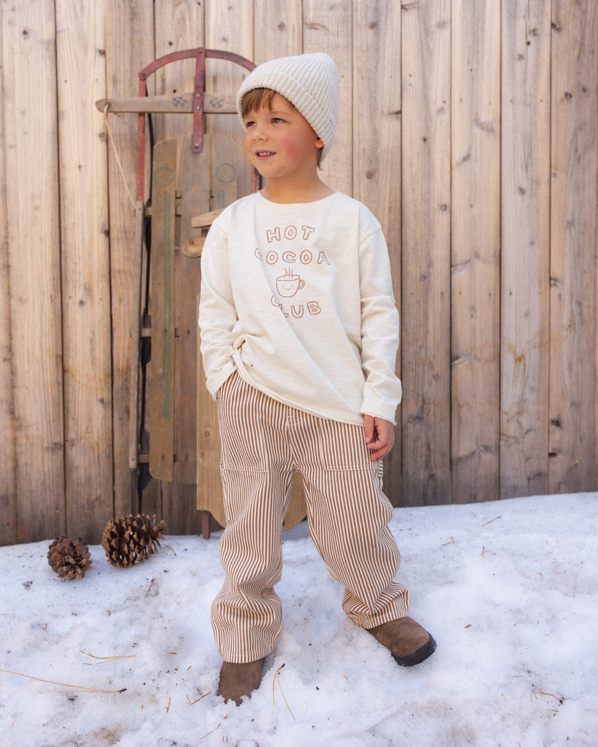 A young child wearing a cream knit beanie, a cream sweatshirt with 'HOT COCOCA CLUB' text, and beige-and-white vertically striped pants, standing outdoors in snow in front of a wooden fence.