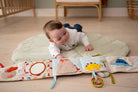 Baby playing with a soft book on a mat in a room with wooden flooring and furniture.
