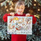 Child holding a colorful illustration of animals in a snowy scene, standing in front of Christmas trees with lights.