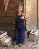 A young boy smiling and wearing blue denim overalls over a red and white striped long-sleeve shirt, standing outdoors in front of a rustic wooden wall with wood logs and a cinderblock nearby.