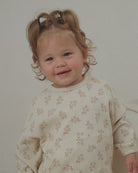 A toddler with curly light brown hair wearing a cream-colored ribbed long-sleeve shirt with a small beige floral pattern, standing against a plain light gray wall.