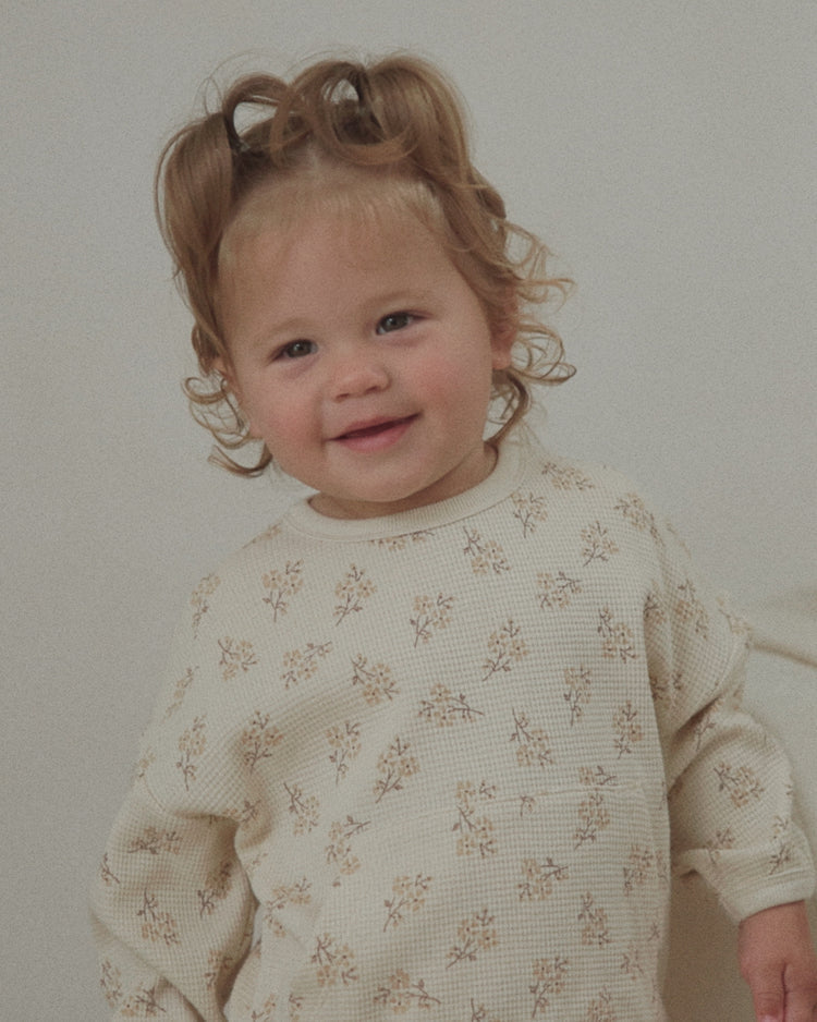 A toddler with curly light brown hair wearing a cream-colored ribbed long-sleeve shirt with a small beige floral pattern, standing against a plain light gray wall.